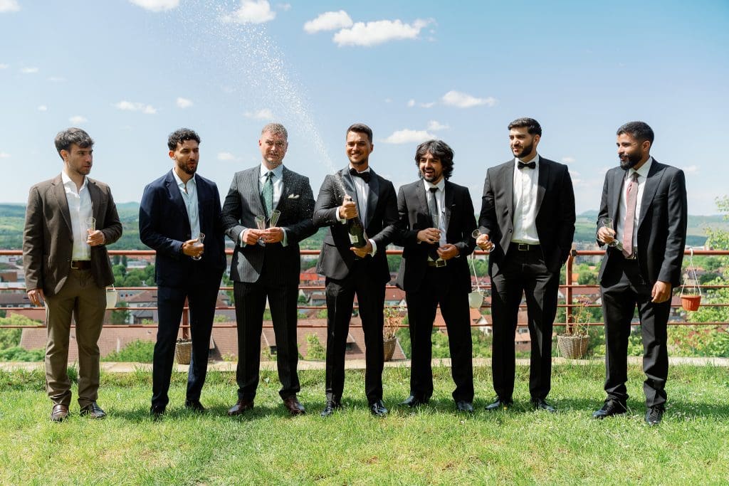 Grup de cavaleri de onoare în costume, deschizând șampanie afară, într-o atmosferă relaxată