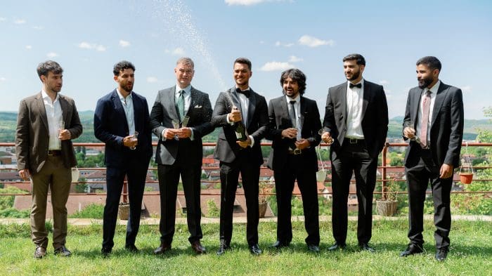 Grup de cavaleri de onoare în costume, deschizând șampanie afară, într-o atmosferă relaxată